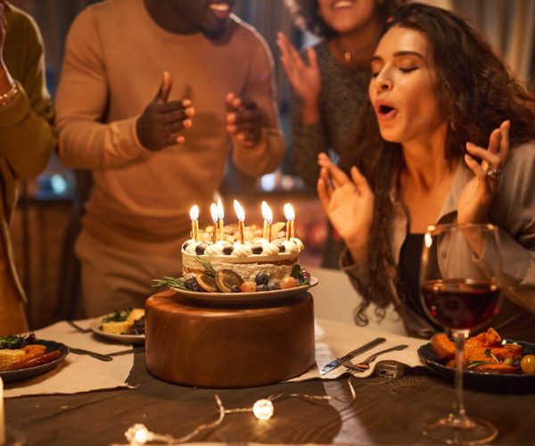 Young woman blowing out candles on birthday cake at the table with her friends clapping hands and congratulating her